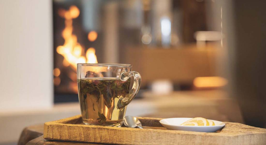 Tee in einer gläsernen Tasse auf einem Holztablett mit Kaminfeuer im Hintergrund als Teil des Heilfasten in Oberstaufen.