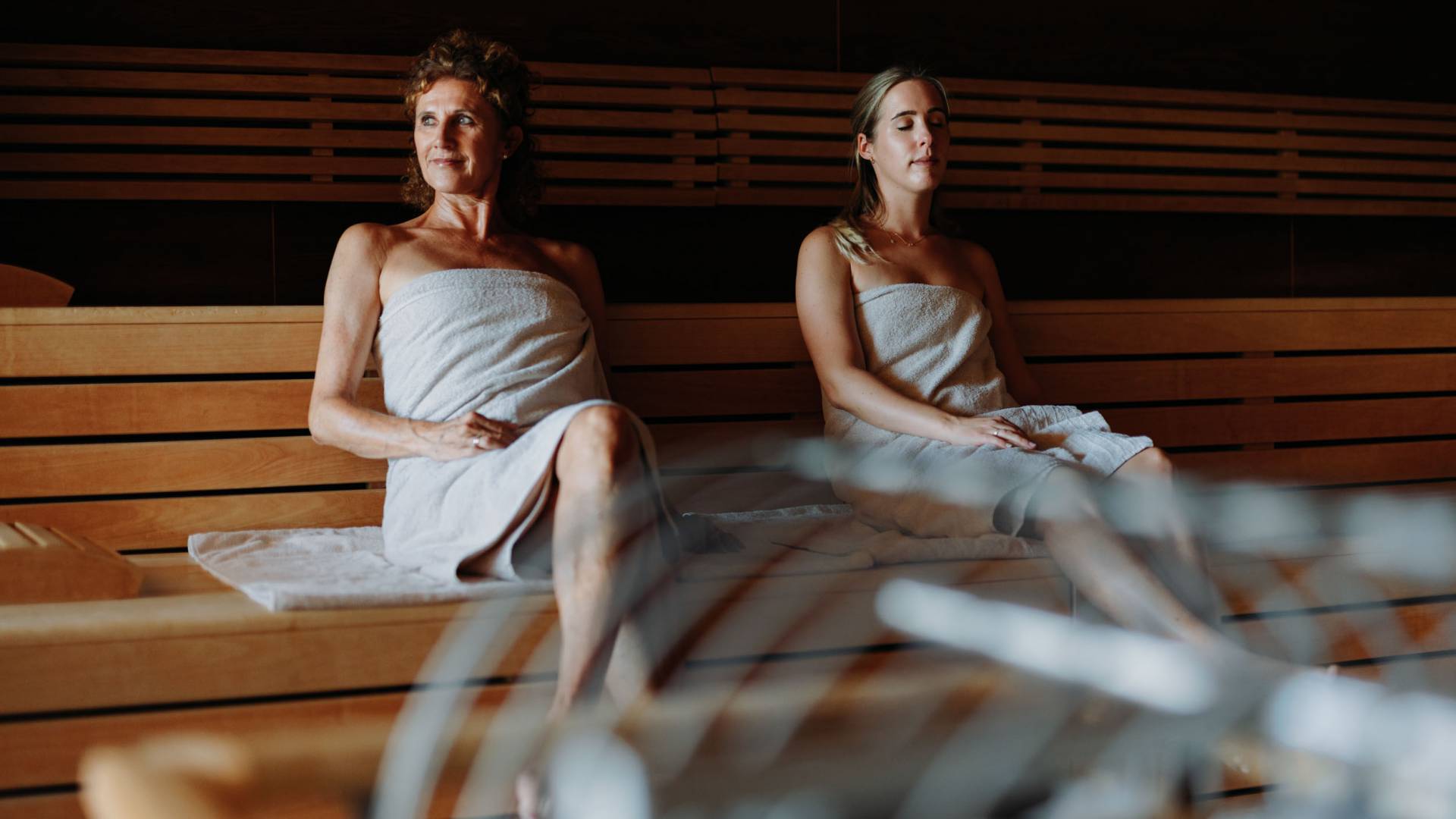 Zwei Frauen sitzen mit hellen Handtüchern nebeneinander in einer Sauna und genießen ihren Winterurlaub in Bayern.