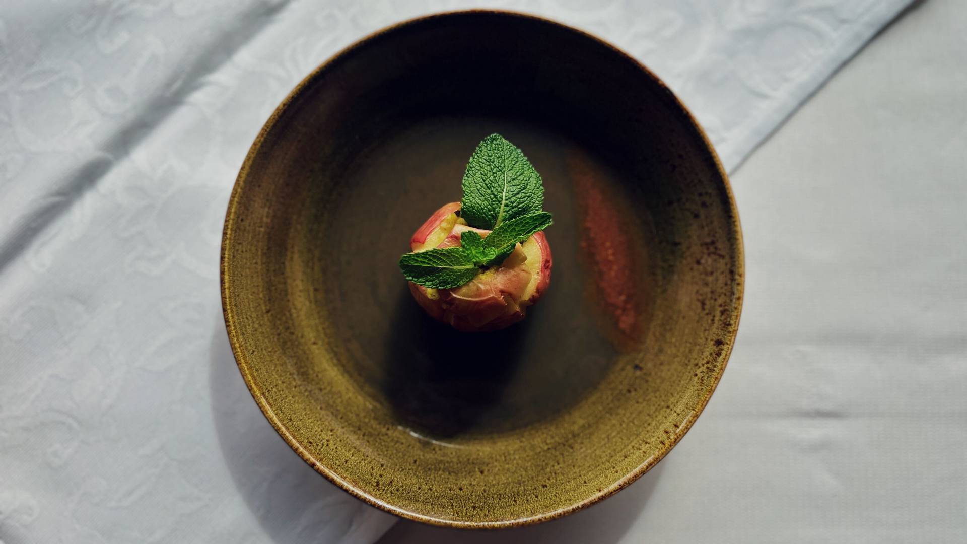 Ein mit Minze verzierter Bratapfel in einem tiefen, naturfarbenem Teller auf einer weißen Tischdecke.