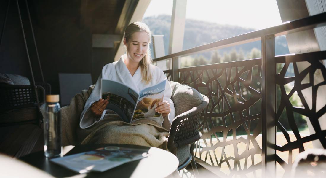 Eine Frau liest ein Magazin auf einem Sessel auf einem Balkon.