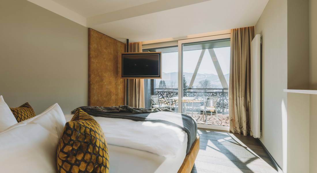 Sicht auf ein warm gestaltetes Hotelzimmer mit Bett, hängendem Flachbildfernseher und großer Glasfront.