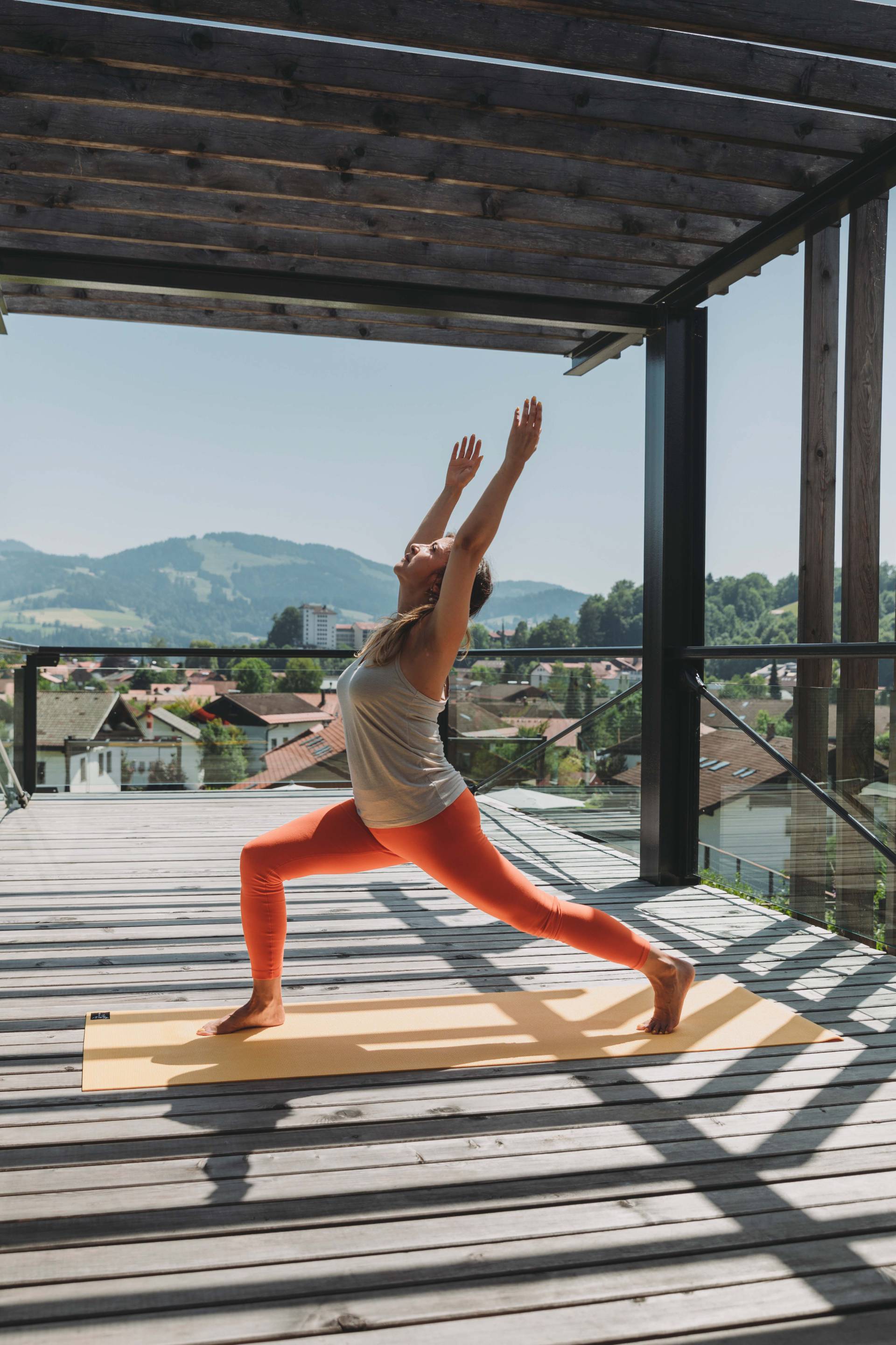 Eine Frau in auffälliger Sporthose macht eine Yogaübung auf einer sonnigen Terrasse als Teil ihres Aktivurlaub im Allgäu.