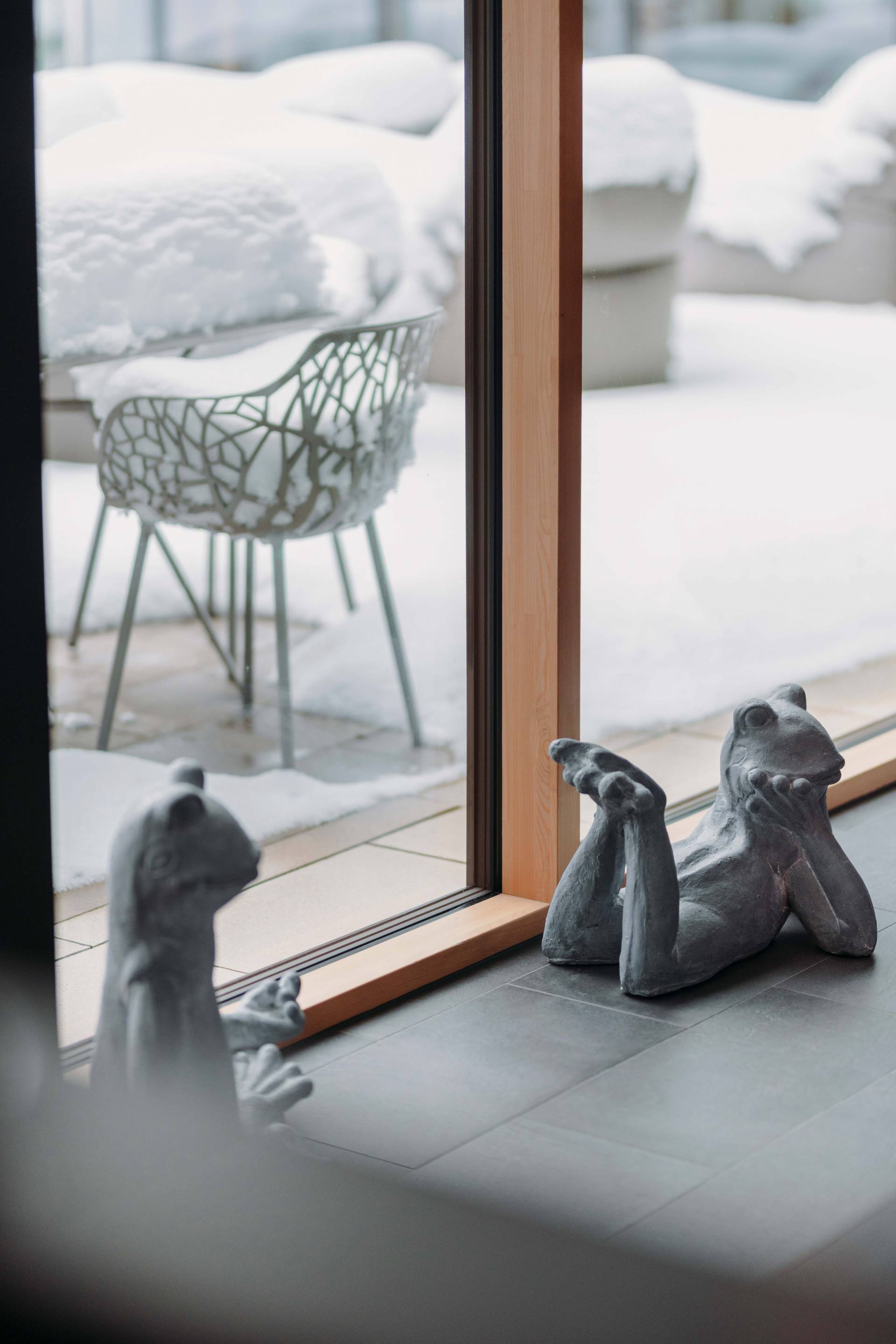 Froschfiguren vor einem Fenster der schneebedeckten Terrasse, welche Lust auf einen Winterurlaub in Bayern macht.