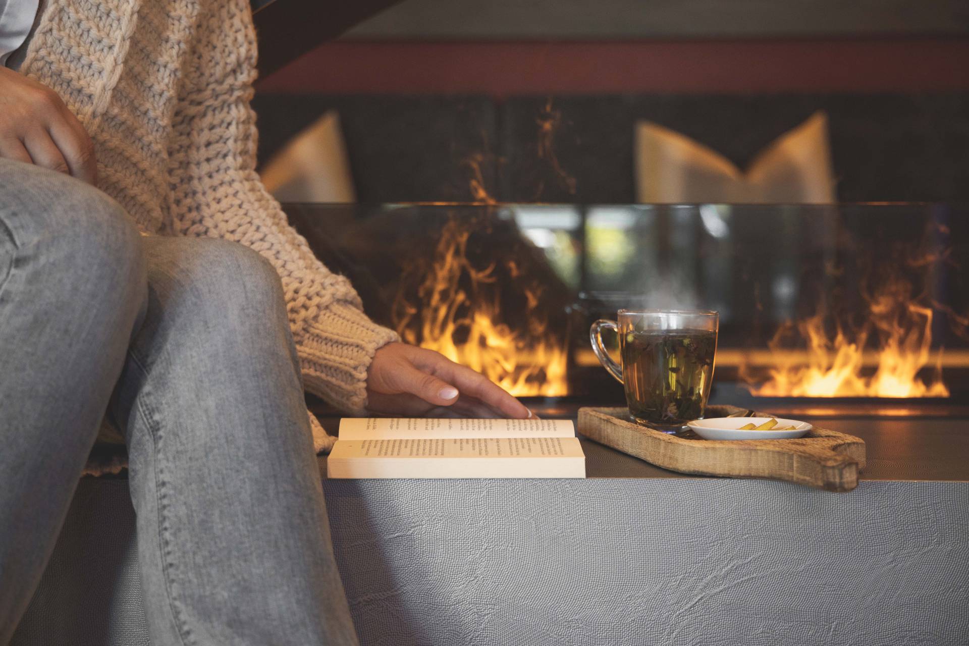 Frau sitzt mit Buch und Tee vor einem Kaminfeuer.