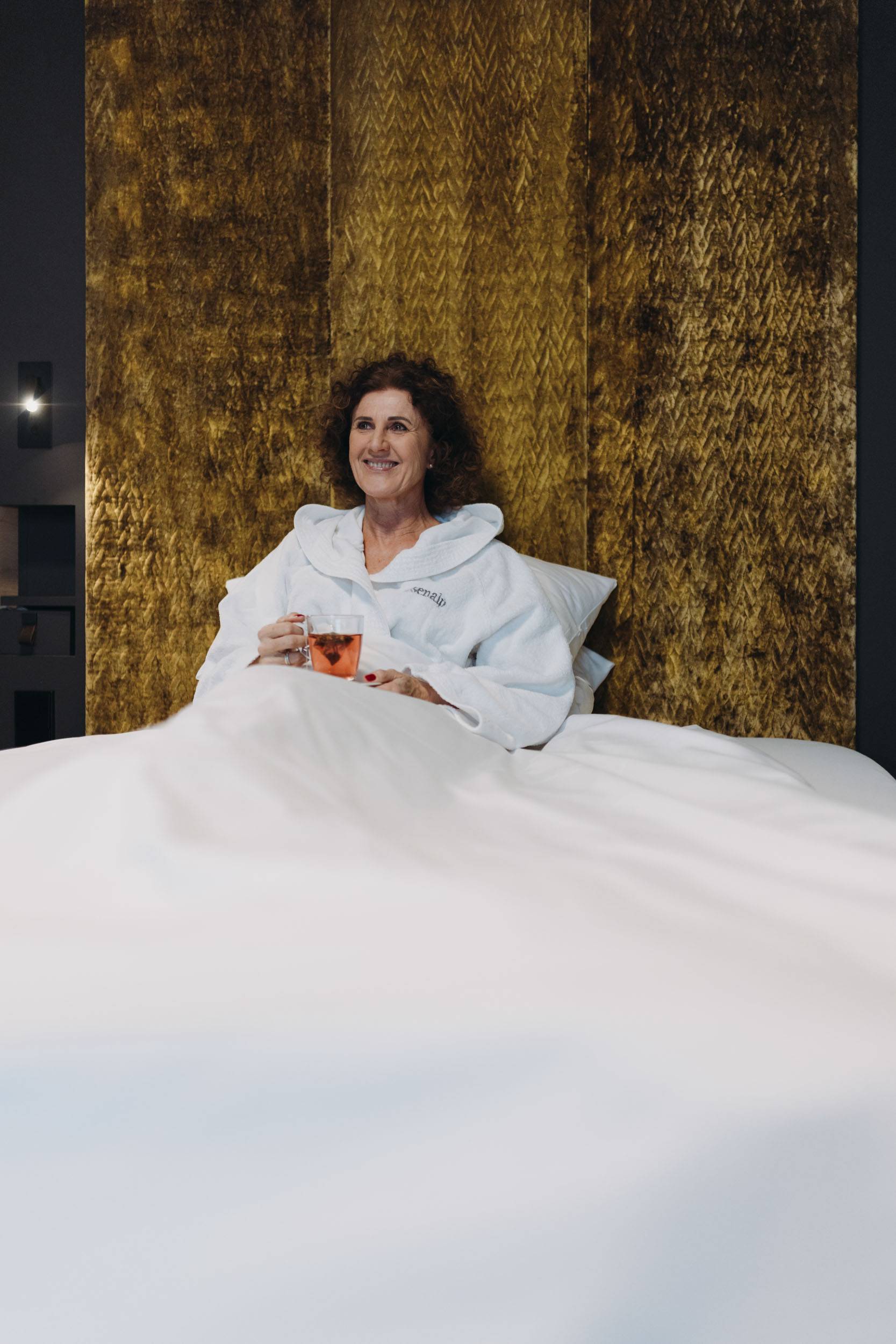 Eine Frau im Bademantel, die entspannt im Bett sitzt mit einer Tasse Tee in der Hand