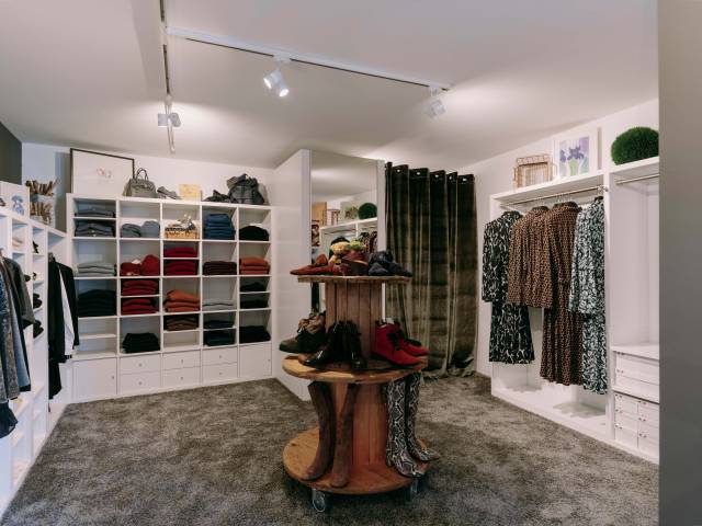 Kleine, stylische Boutique mit Klamotten in weißen Wandregalen und Schuhen auf einem Holzmöbel präsentiert.