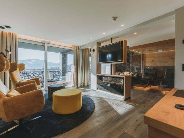 Eine gemütliche Wellness-Suite mit warmen Farben, zwei Sesseln, Fernseher und einem gläsernen Saunabereich.