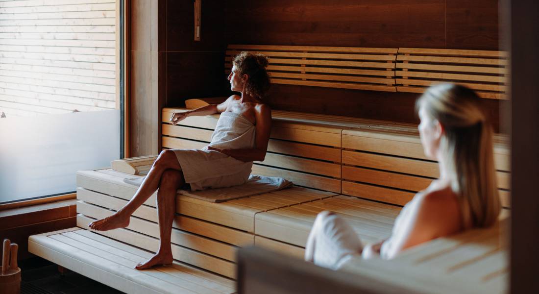 Zwei Frauen sitzen mit weißen Handtüchern in einer Sauna.
