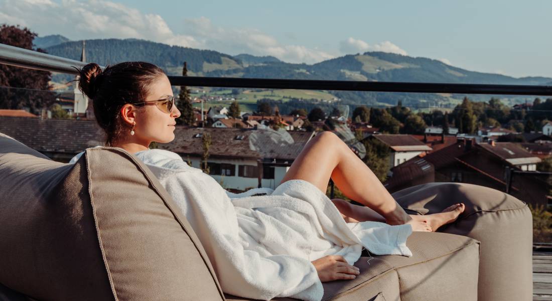 Eine Frau im Bademantel entspannt auf der Terrasse mit Bergpanorama.
