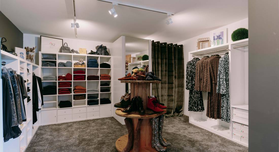Kleine, stylische Boutique mit Klamotten in weißen Wandregalen und Schuhen auf einem Holzmöbel präsentiert.