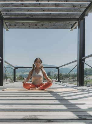 Frau sitzt im Schneidersitz auf Yogamatte auf einer Terrasse.