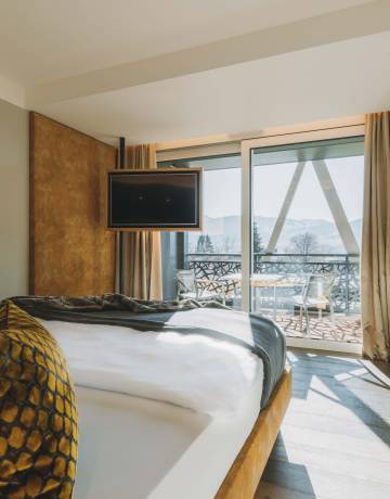 Sicht auf ein warm gestaltetes Hotelzimmer mit Bett, hängendem Flachbildfernseher und großer Glasfront.