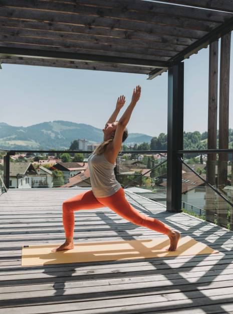Eine Frau in auffälliger Sporthose macht eine Yogaübung auf einer sonnigen Terrasse als Teil ihres Aktivurlaub im Allgäu.