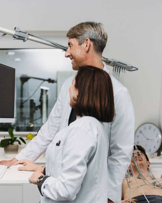 Eine Ärztin und ein Arzt stehen an einem Computer mit einem Patienten, welcher im Hintergrund liegt.