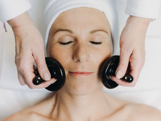 Eine Frau mit geschlossenen Augen erhält auf dem Rücken liegend eine Gesichtsbehandlung mit zwei Massageinstrumenten.