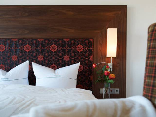 Sicht auf ein weiß bezogenes Bett mit einer Rückenlehne mit bunter Verzierung und Blumen auf dem Nachttisch.