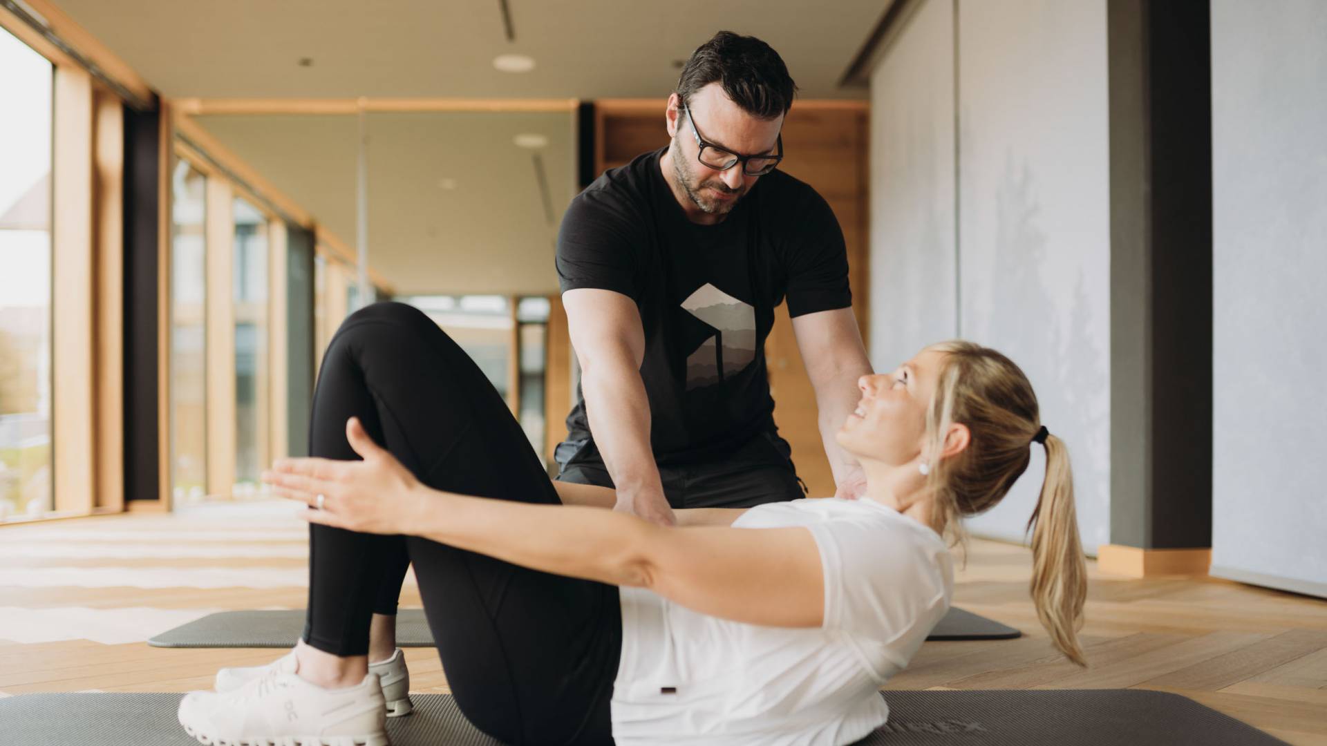 Eine junge Frau erhält Hilfestellung bei einer Fitnessübung auf einer Gymnastikmatte in einem modernen Studio.