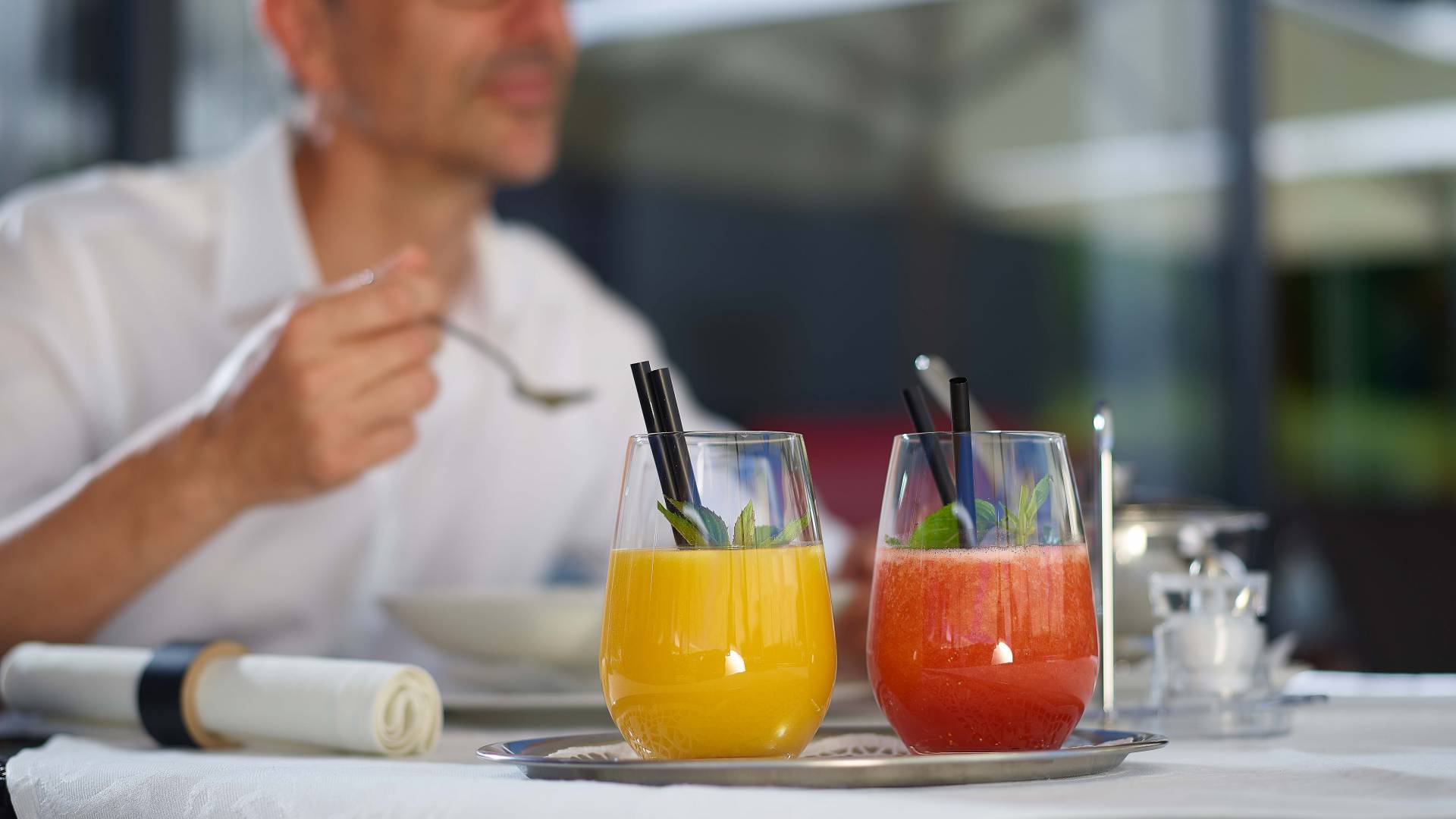 Zwei Gläser mit unterschiedlichen Säften als Teil des Heilfasten in Oberstaufen auf einem silbernen Tablett und einem Restaurantgast im Hintergrund.