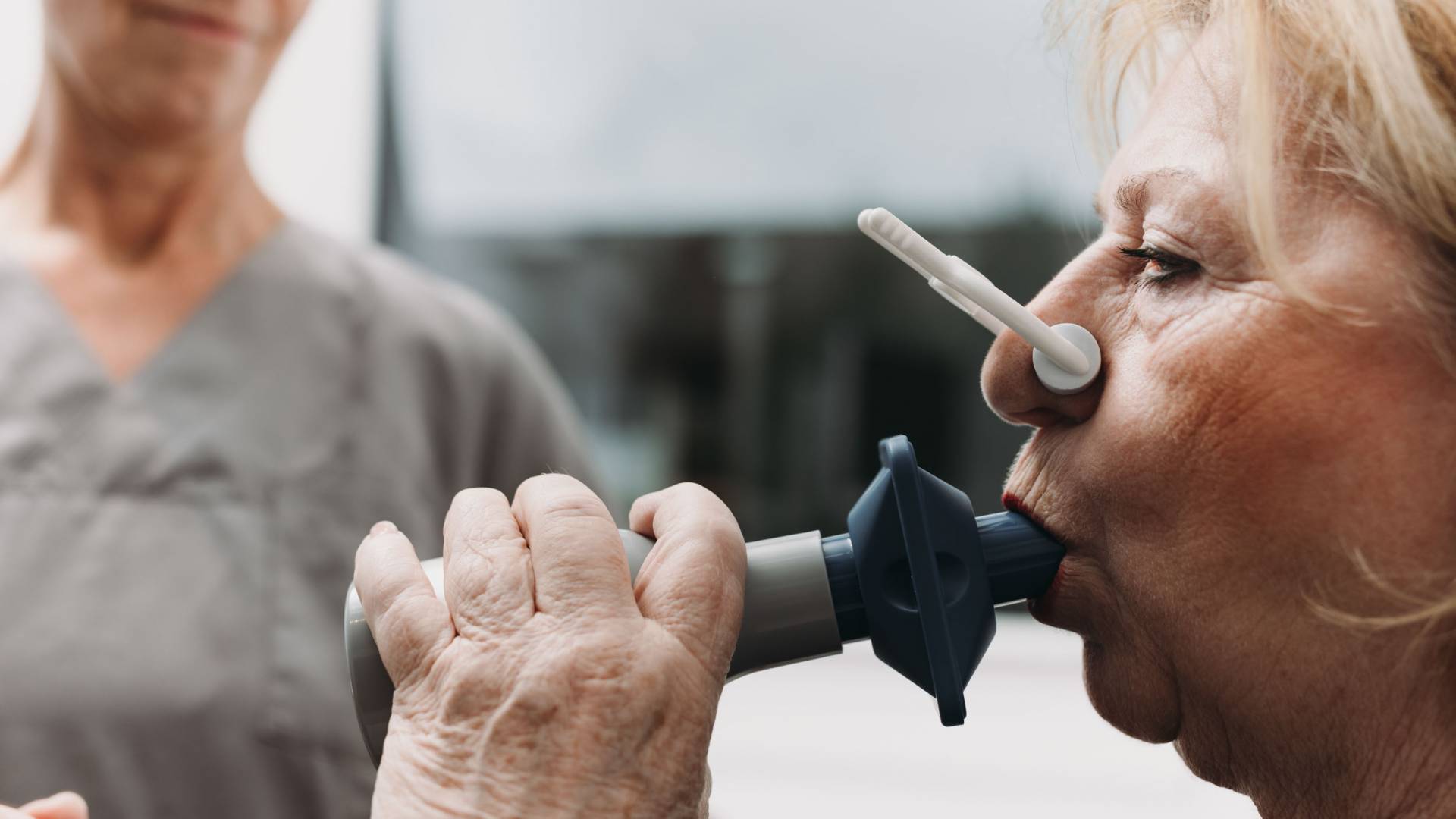 Eine ältere Frau mit Nasenklemme pustet in ein Messgerät.