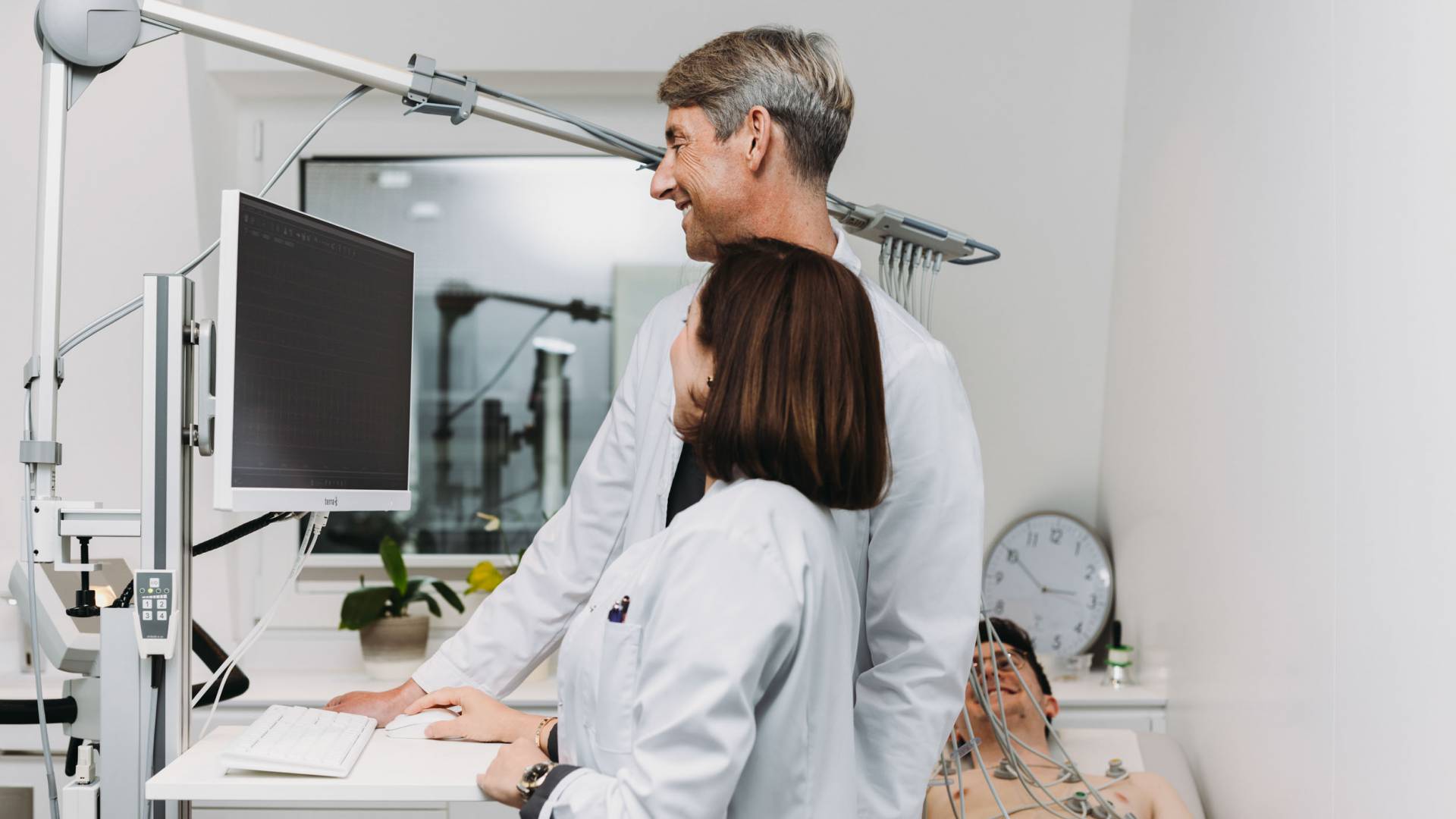 Eine Ärztin und ein Arzt stehen an einem Computer mit einem Patienten, welcher im Hintergrund liegt.