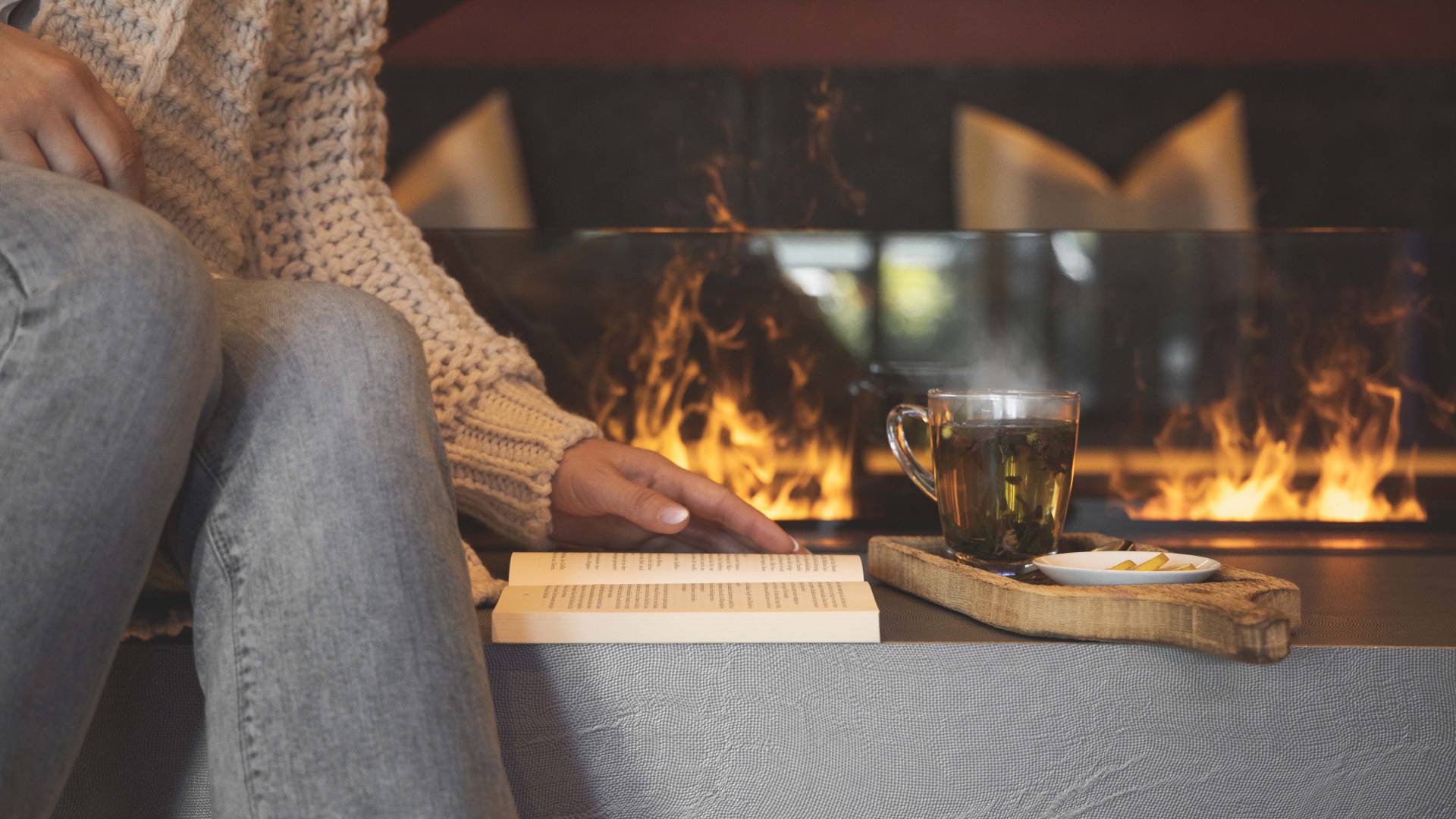 Frau sitzt mit Buch und Tee vor einem Kaminfeuer.