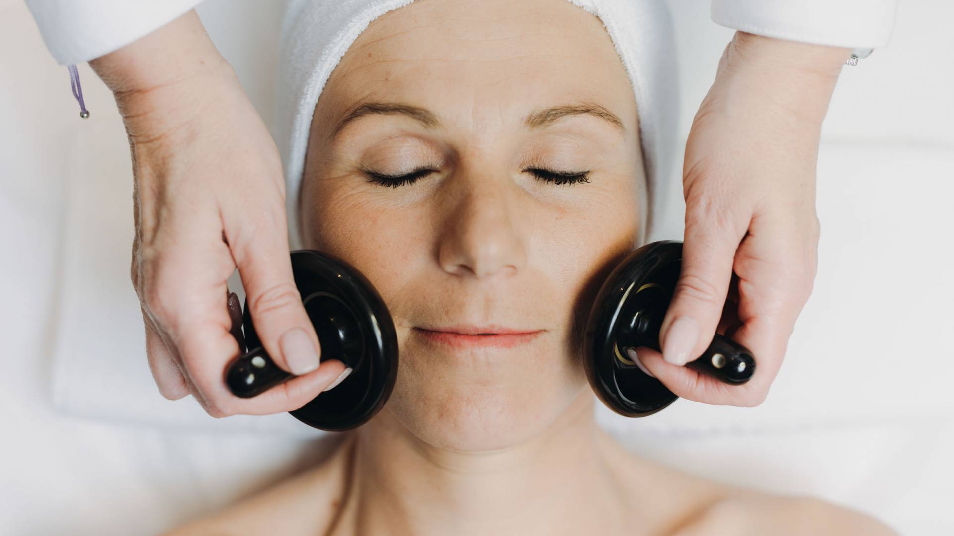 Eine Frau mit geschlossenen Augen erhält auf dem Rücken liegend eine Gesichtsbehandlung mit zwei Massageinstrumenten.